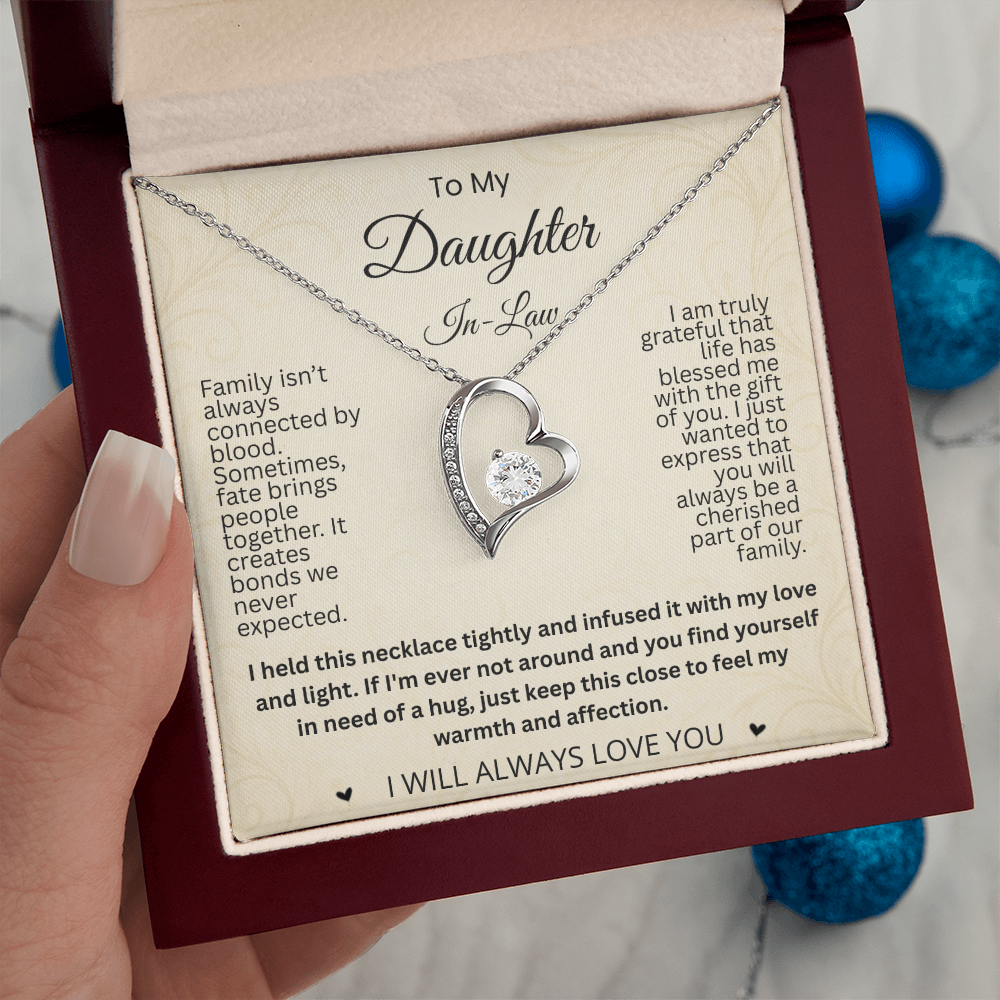 Hand holding a reddish-brown gift box with a silver-tone heart necklace and daughter-in-law card above blue ornaments.