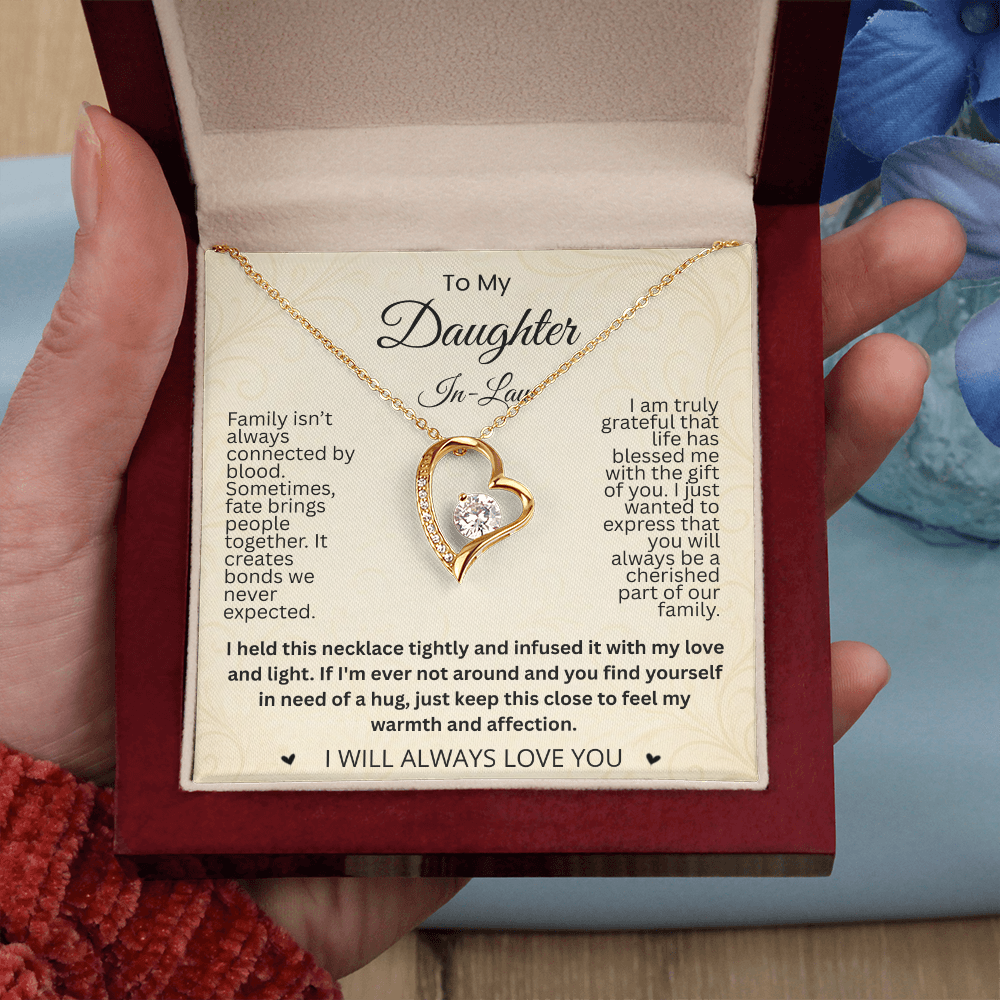 Hand holding a reddish-brown gift box with a gold-tone heart necklace and daughter-in-law card beside blue flowers.