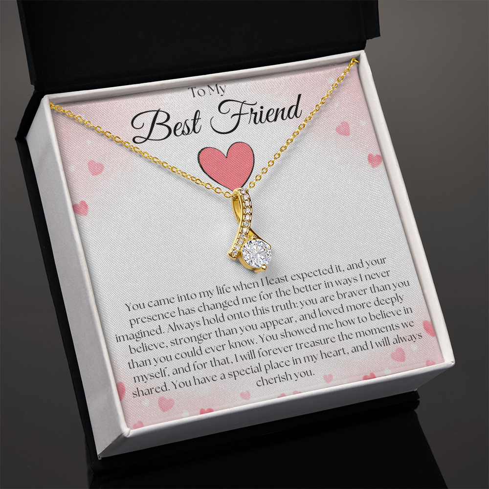 Angled view of a gold-tone pendant necklace and pink-heart best-friend card in a black and white gift box on black.