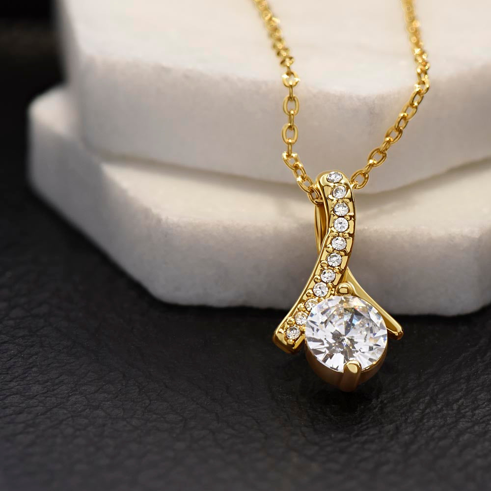 Close-up of a gold-tone curved pendant with a round clear stone beside a white display block on black.