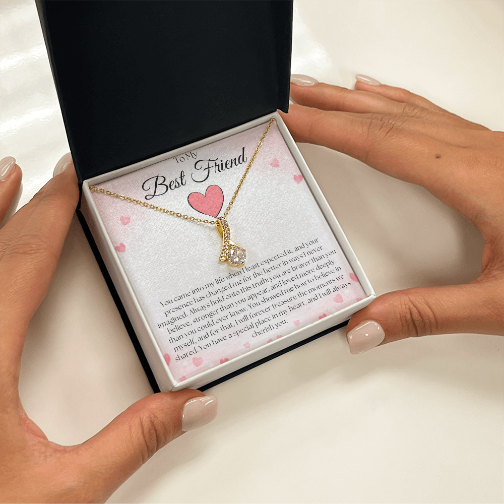 Hands framing a black and white gift box with a gold-tone pendant necklace and pink-heart best-friend card on white.