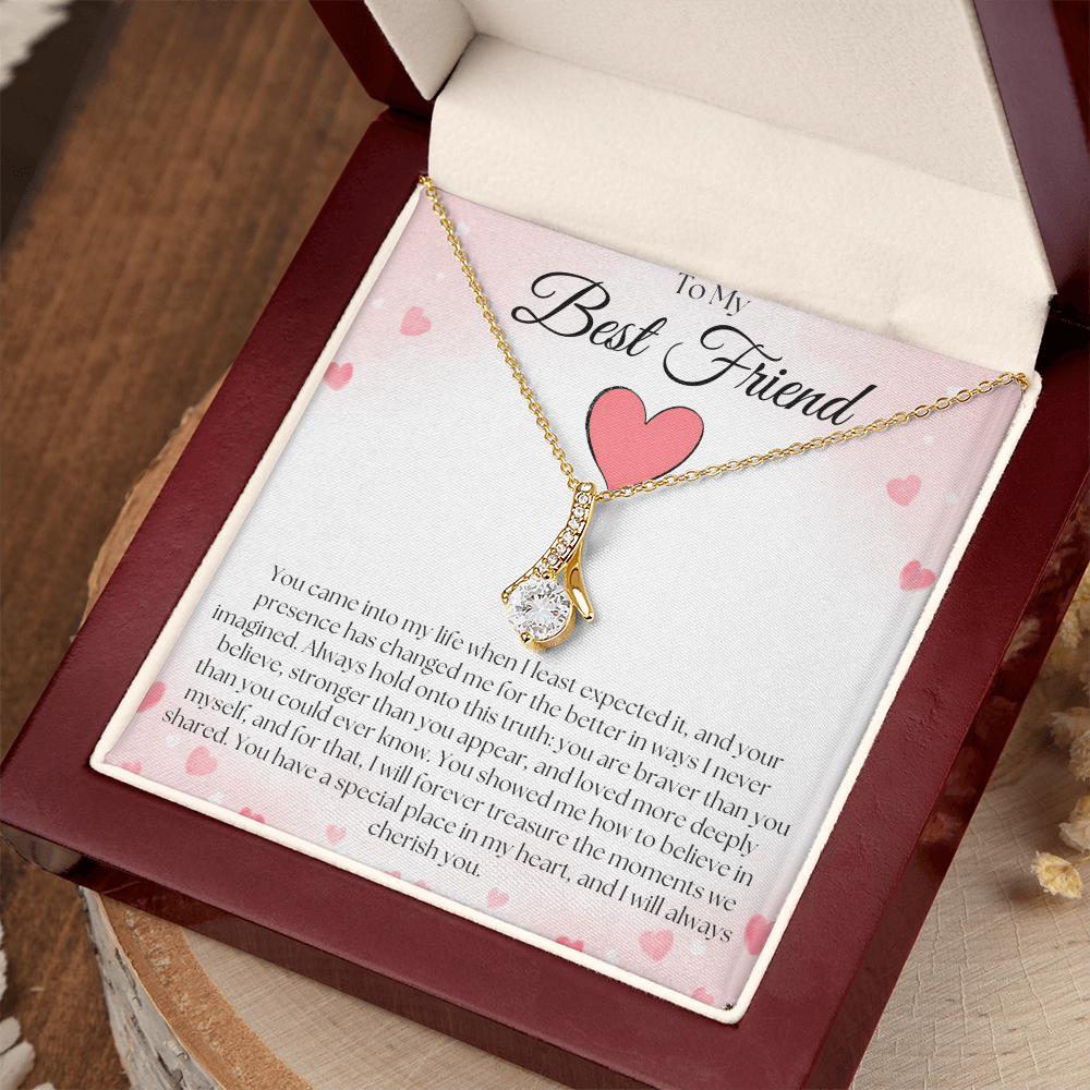 Gold-tone pendant necklace and pink-heart best-friend card in a reddish-brown gift box on a wood slice.