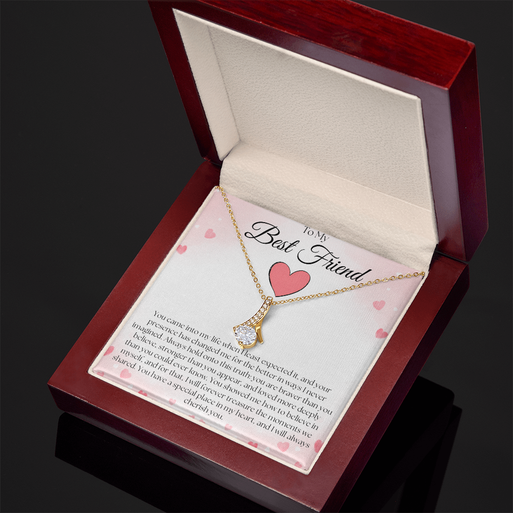 Angled view of a gold-tone pendant necklace and best-friend card in a reddish-brown gift box on a dark reflective surface.