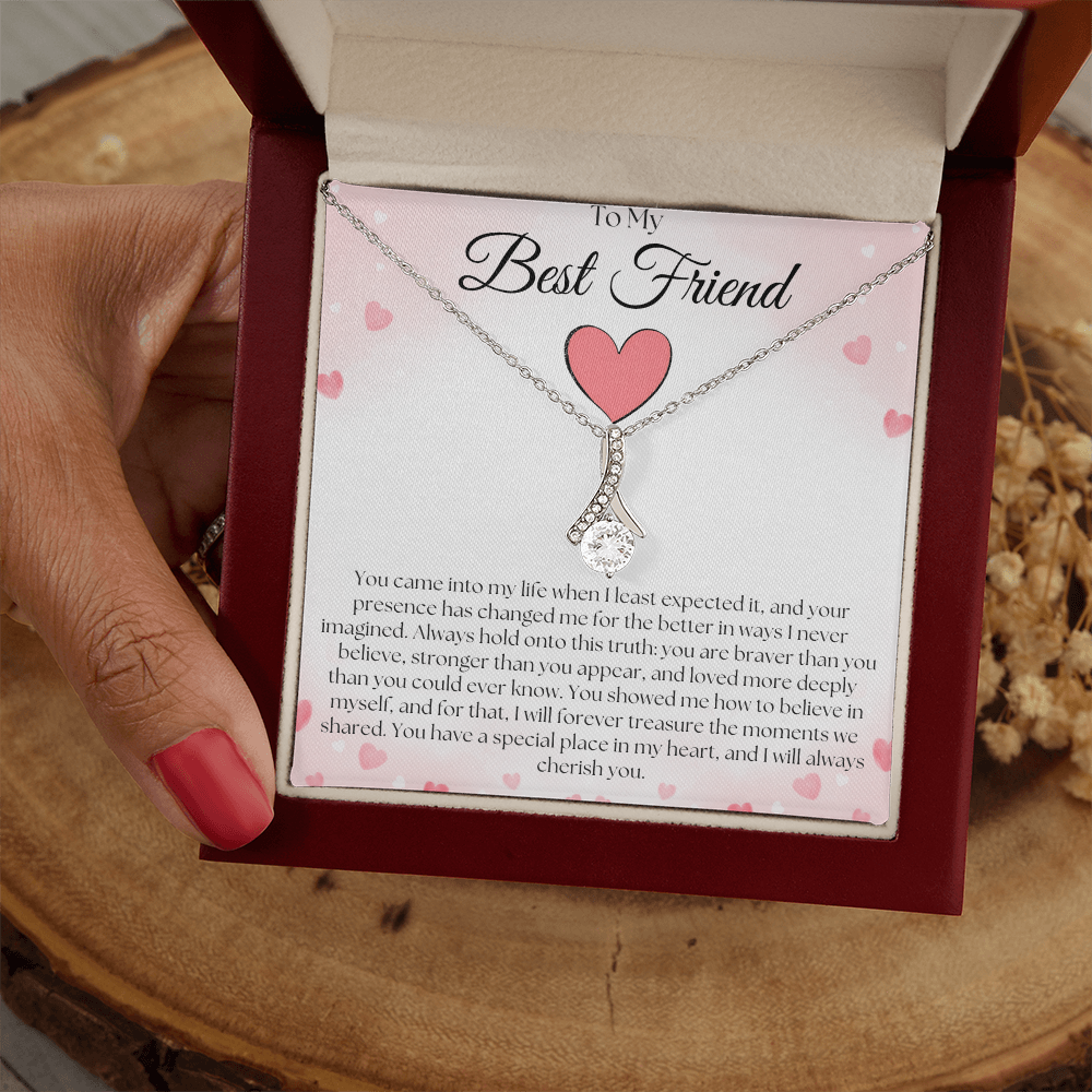 Hand holding a reddish-brown gift box with a silver-tone pendant necklace and best-friend card over a wood slice.