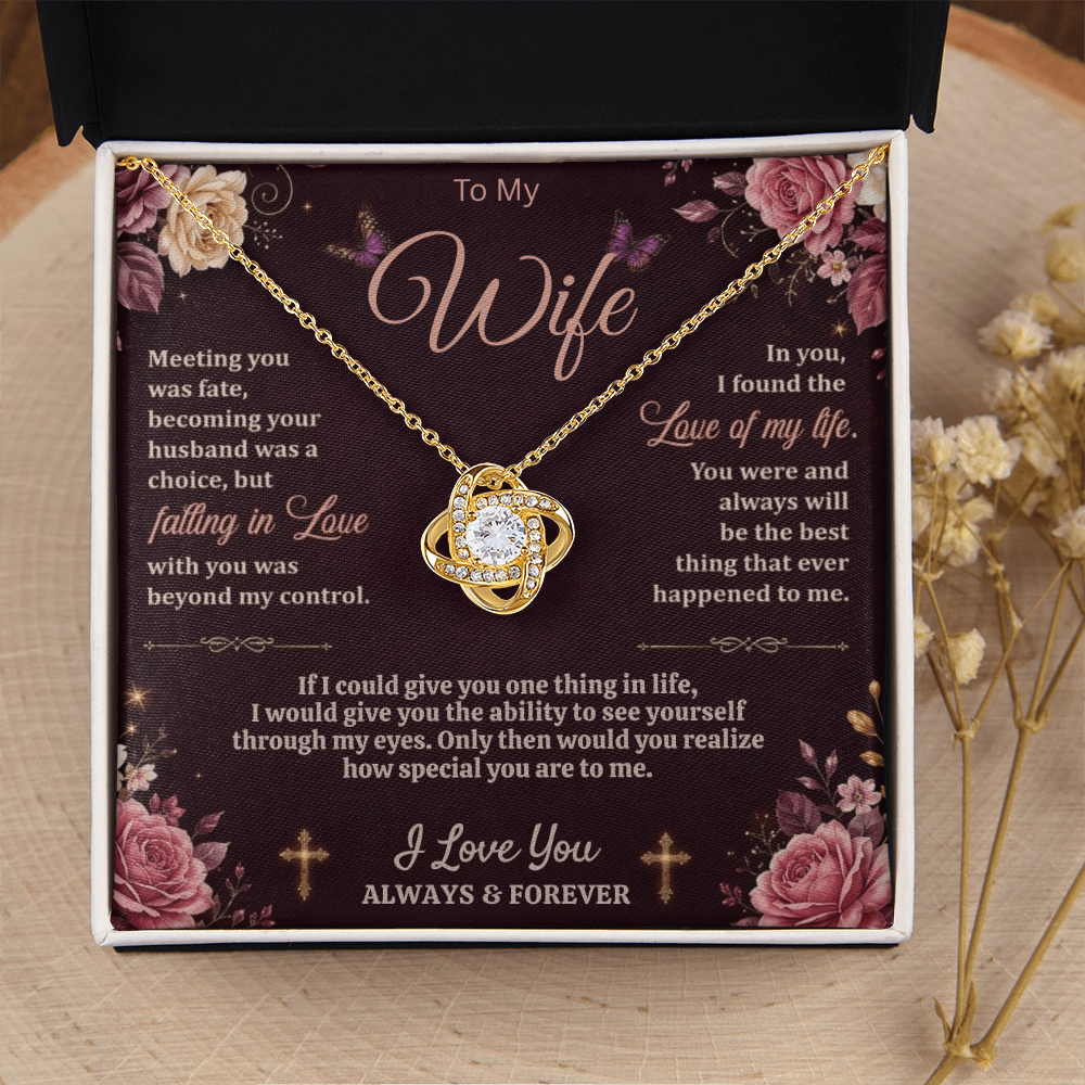 Gold-tone love-knot necklace and dark floral wife card in a black and white gift box on a wood slice beside dried flowers.