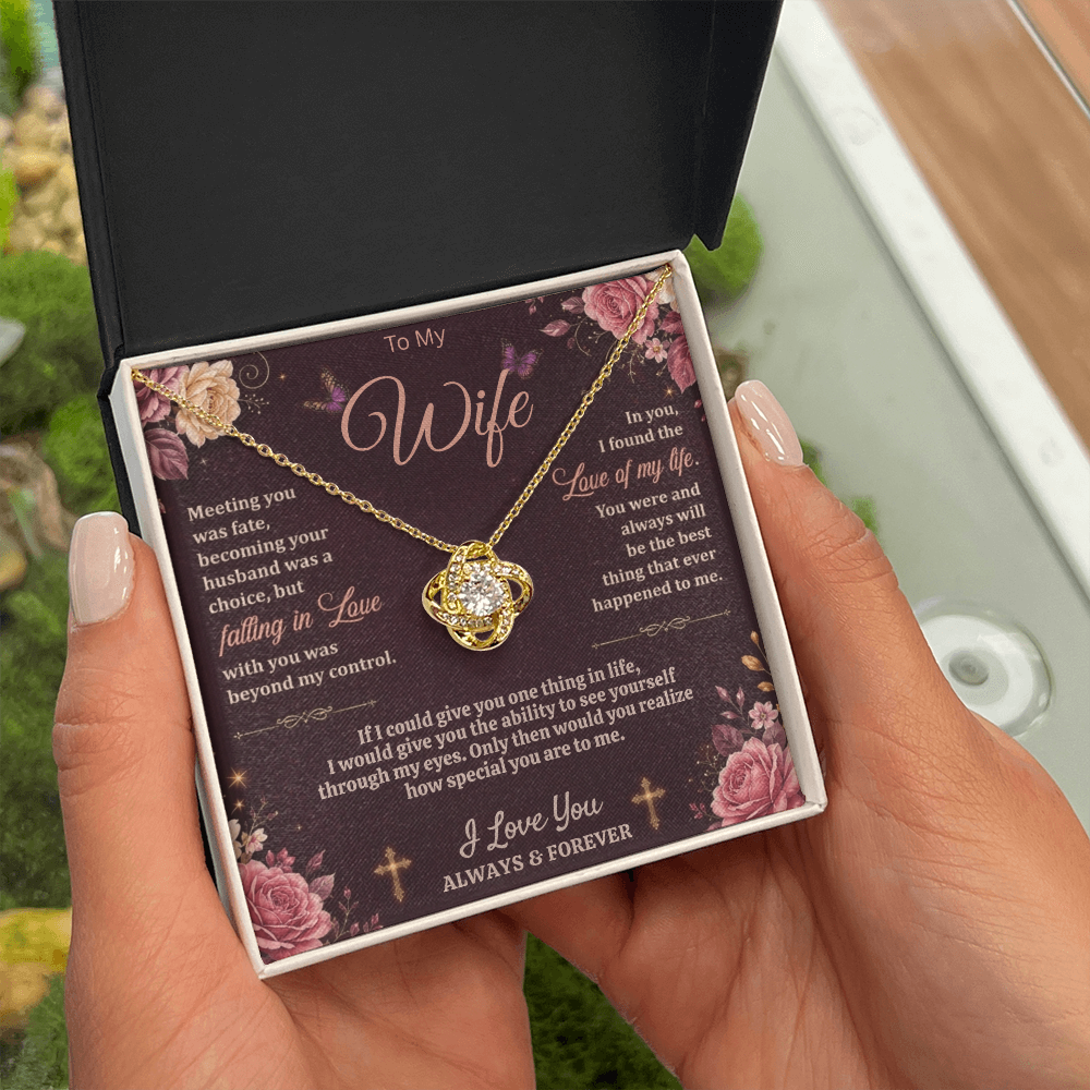 Hands holding a black and white gift box with a gold-tone love-knot necklace and dark floral wife message card.
