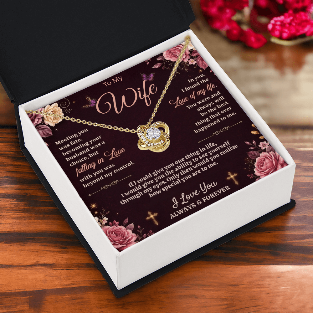 Gold-tone love-knot necklace and dark floral wife card in a black and white gift box on wood beside red decorations.