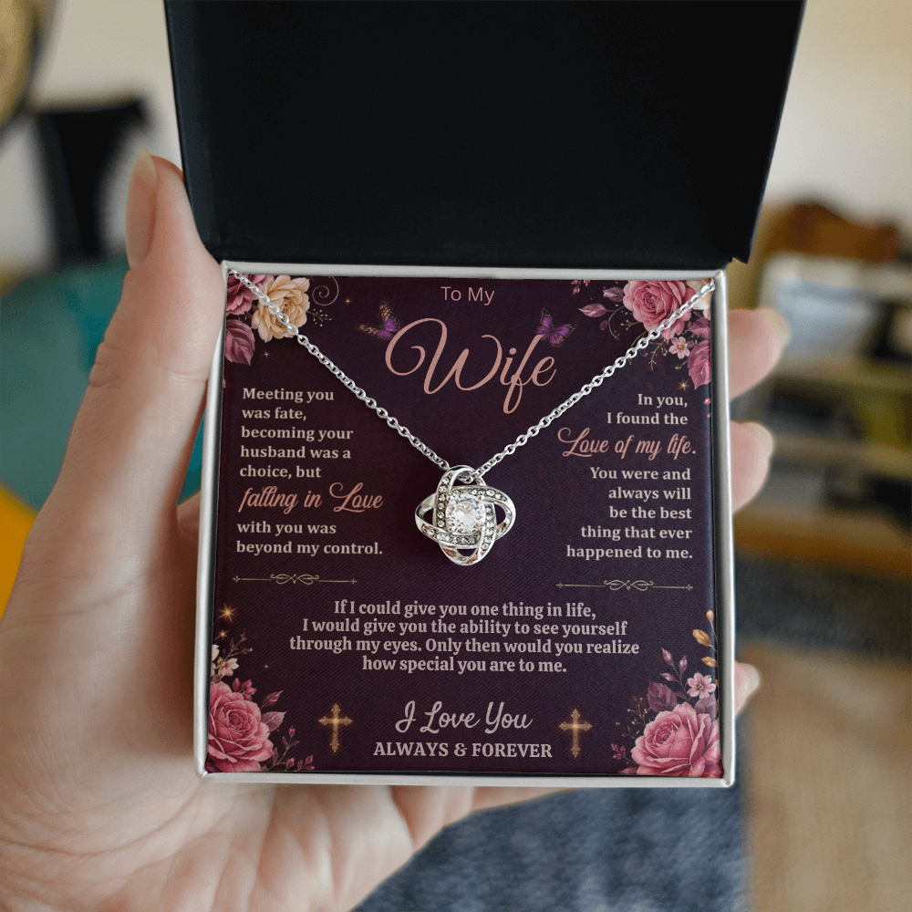 Hand holding a black and white gift box with a silver-tone love-knot necklace and dark floral wife message card indoors.