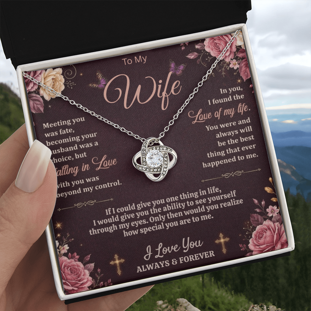 Hand holding a black and white gift box with a silver-tone love-knot necklace and dark floral wife card against a mountain landscape.
