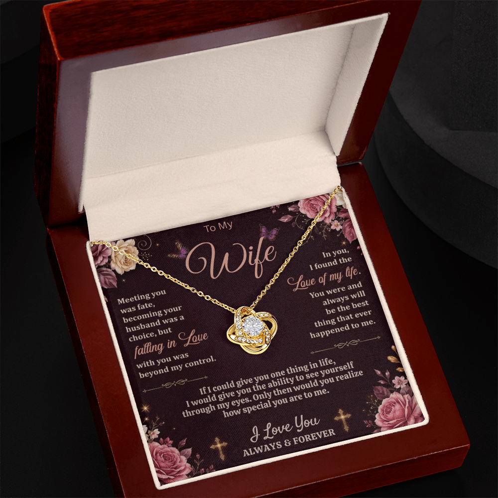 Angled view of a gold-tone love-knot necklace and dark floral wife card in a reddish-brown gift box on black.