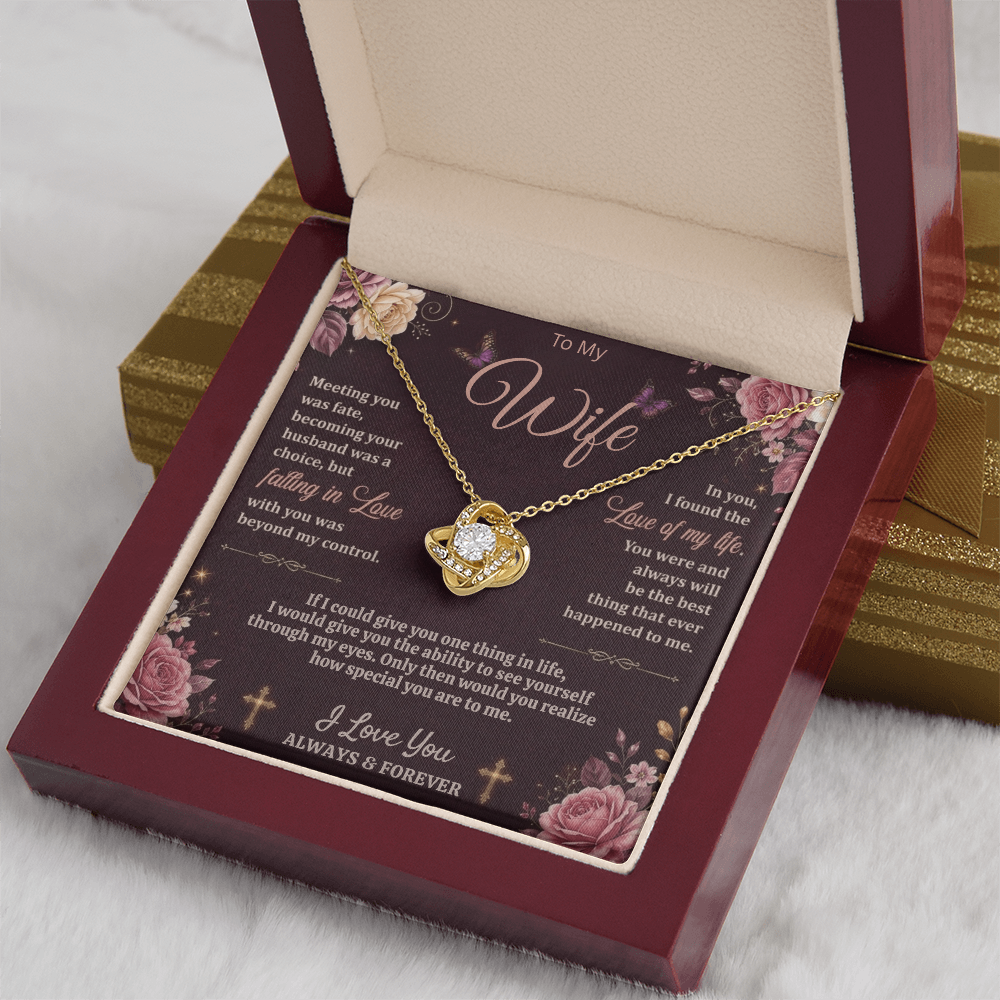 Gold-tone love-knot necklace and dark floral wife card in a reddish-brown gift box on white fabric beside a gold gift box.