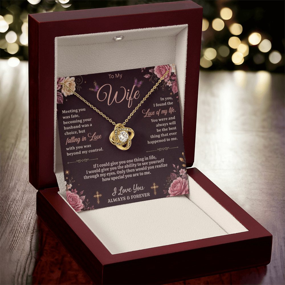 Gold-tone love-knot necklace and dark floral wife card displayed upright in a reddish-brown gift box on wood with blurred lights behind.