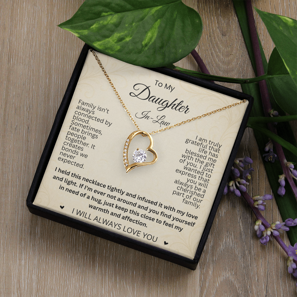 Gold-tone heart necklace and daughter-in-law card in a black gift box on wood beside green leaves and purple flowers.